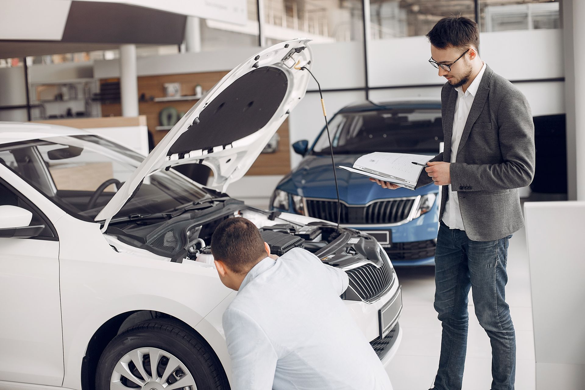 Man examines car engine while another reviews paperwork at a dealership.