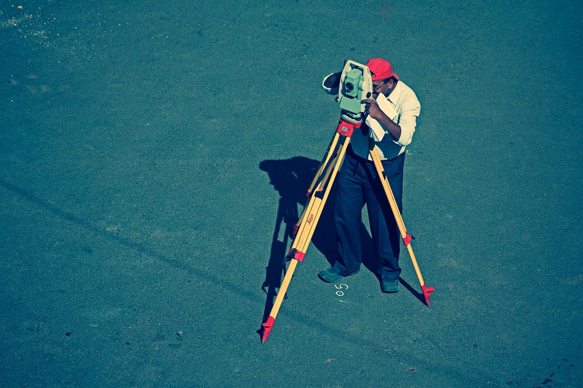 Surveyor in a red cap uses surveying equipment on a tripod outdoors; casting a shadow on a light gray surface.