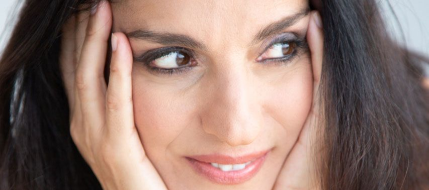 Γυναίκα με σκούρα μαλλιά, τα χέρια στο πρόσωπό της, κοιτάζει στο πλάι με ένα απαλό χαμόγελο.