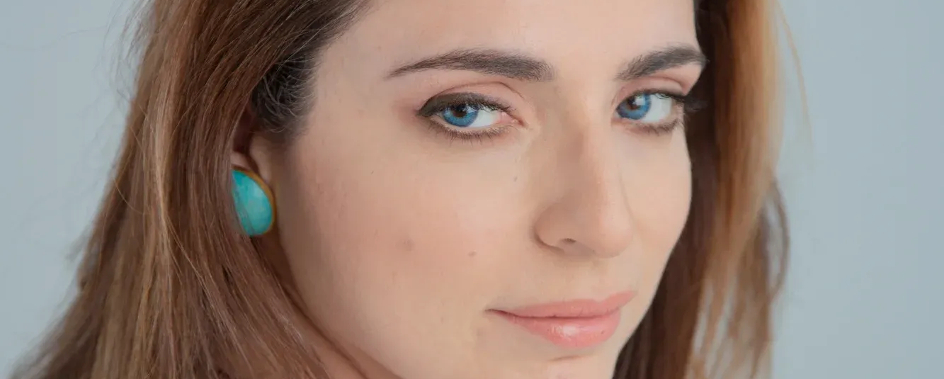 Γυναίκα με σκούρα μαλλιά, τα χέρια στο πρόσωπό της, κοιτάζει στο πλάι με ένα απαλό χαμόγελο.