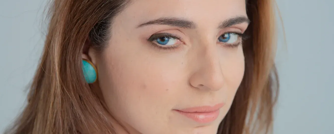 Γυναίκα με σκούρα μαλλιά, τα χέρια στο πρόσωπό της, κοιτάζει στο πλάι με ένα απαλό χαμόγελο.