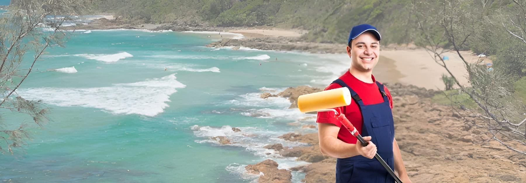 A Man is Holding a Paint Roller in Front of a Beach — O'Brien Painting Contractors in Alexandra Headland, QLD