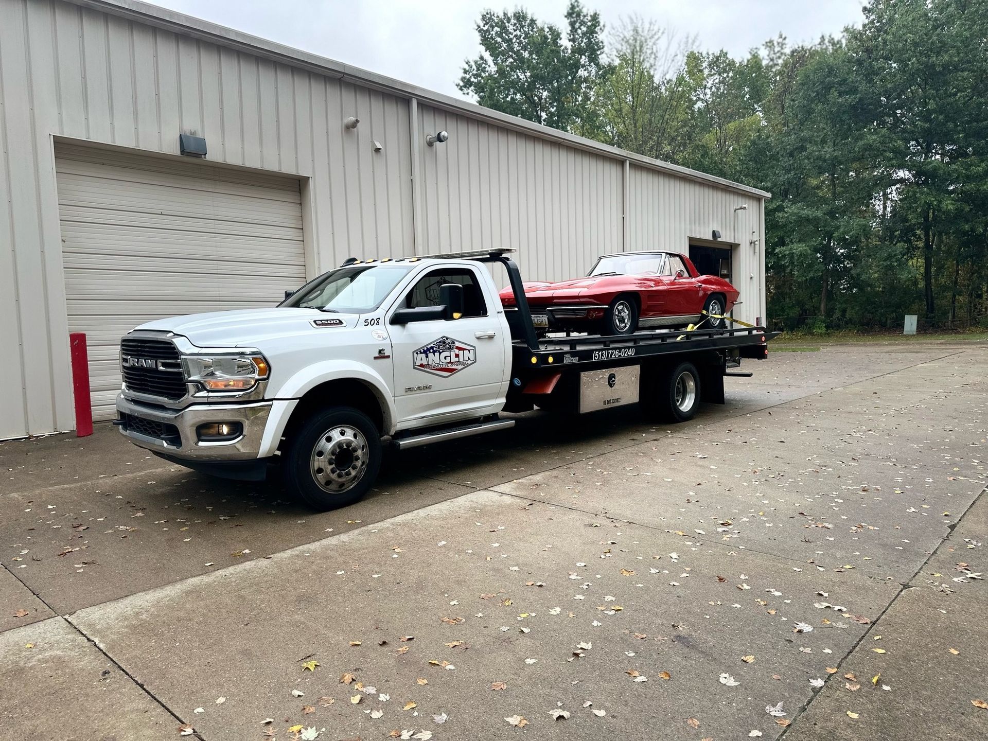 White tow truck carrying a red classic car, parked in front of a building with a closed garage door.