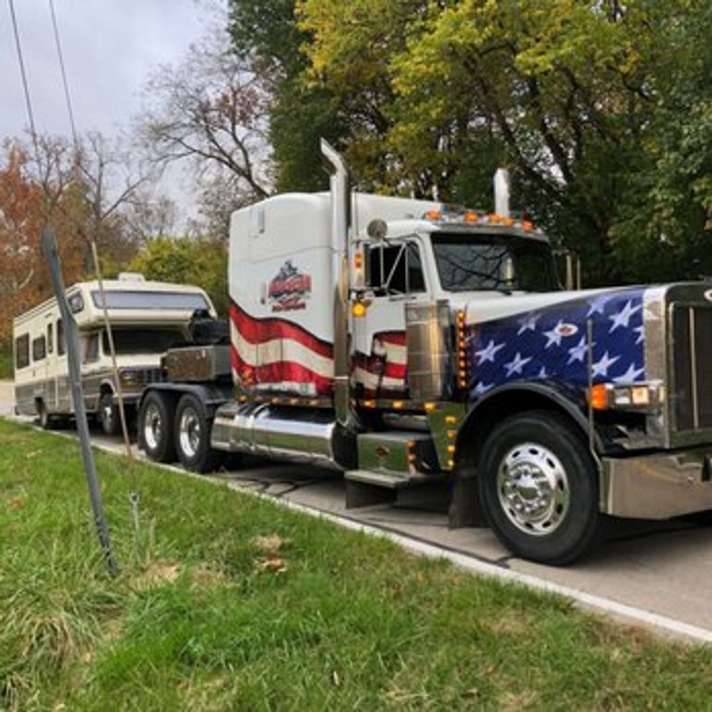 Semi-truck with American flag design towing a camper on a road, trees in the background.