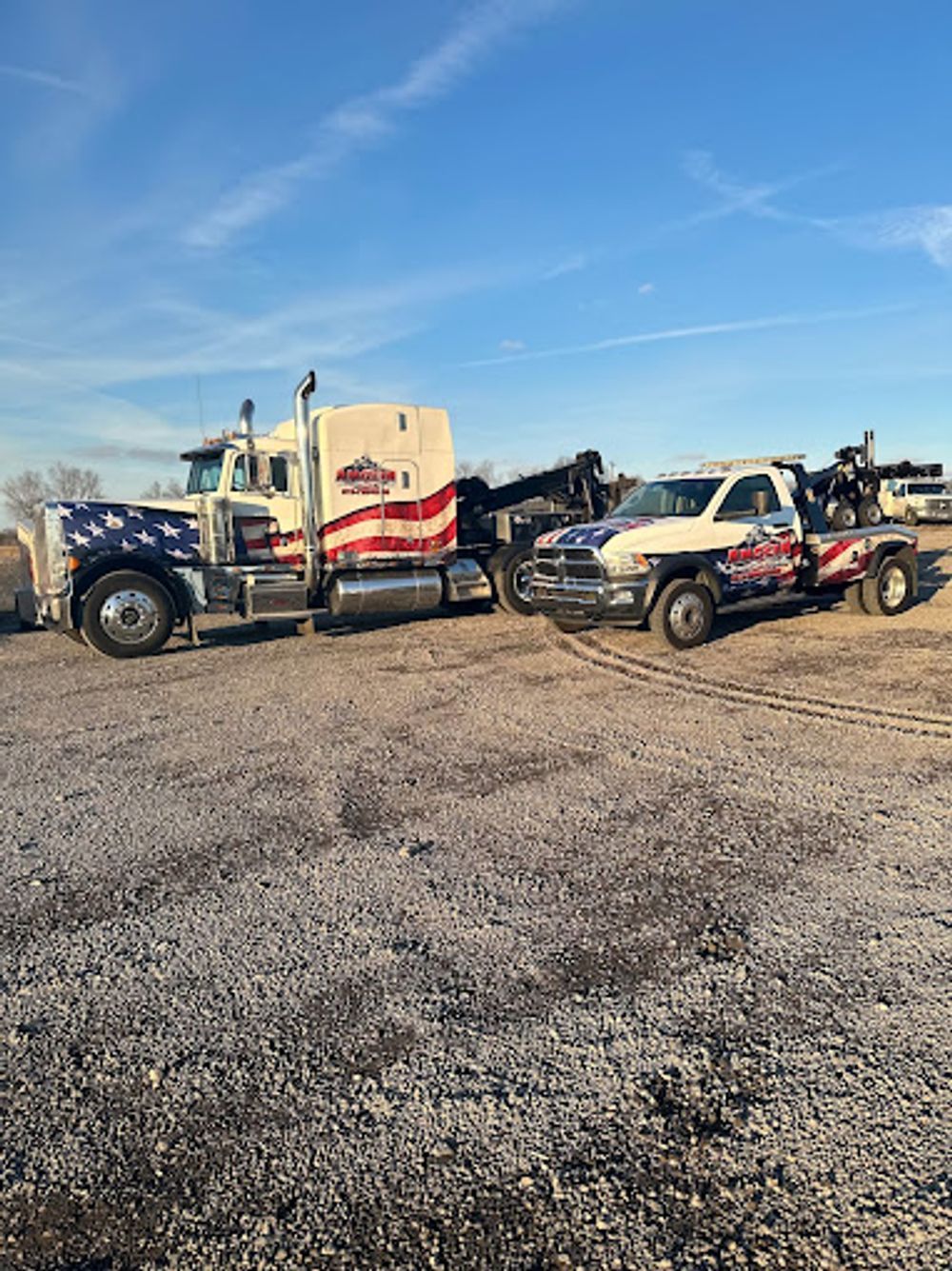 Two tow trucks with American flag designs parked on gravel under a blue sky.