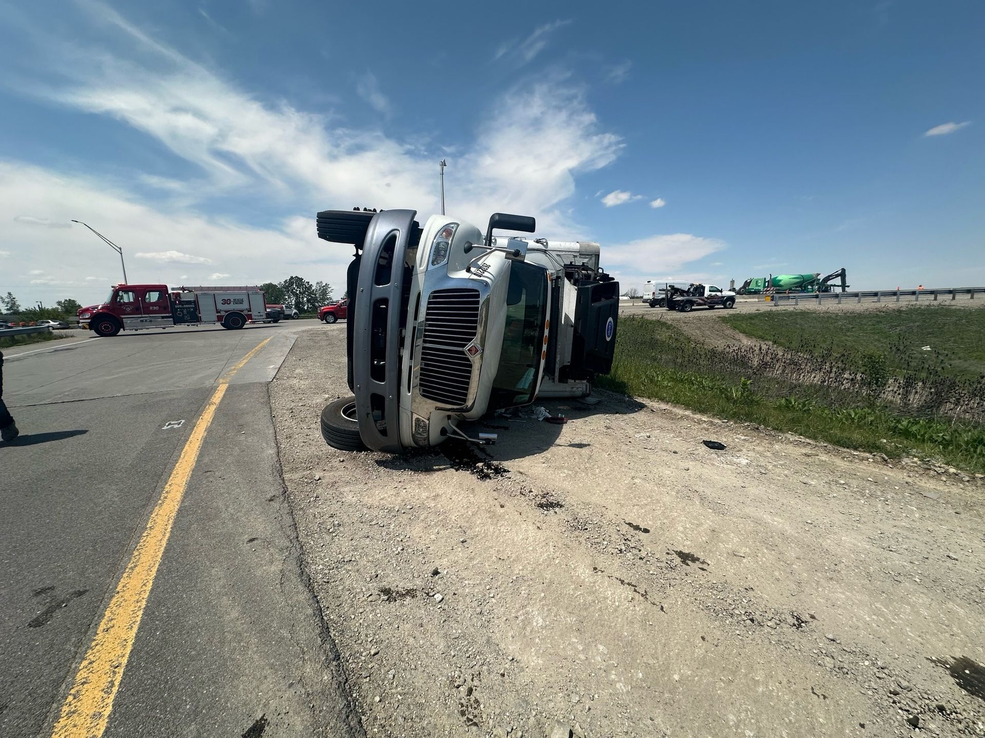 A semi-truck overturned on a gravel shoulder of a highway, under a bright blue sky.