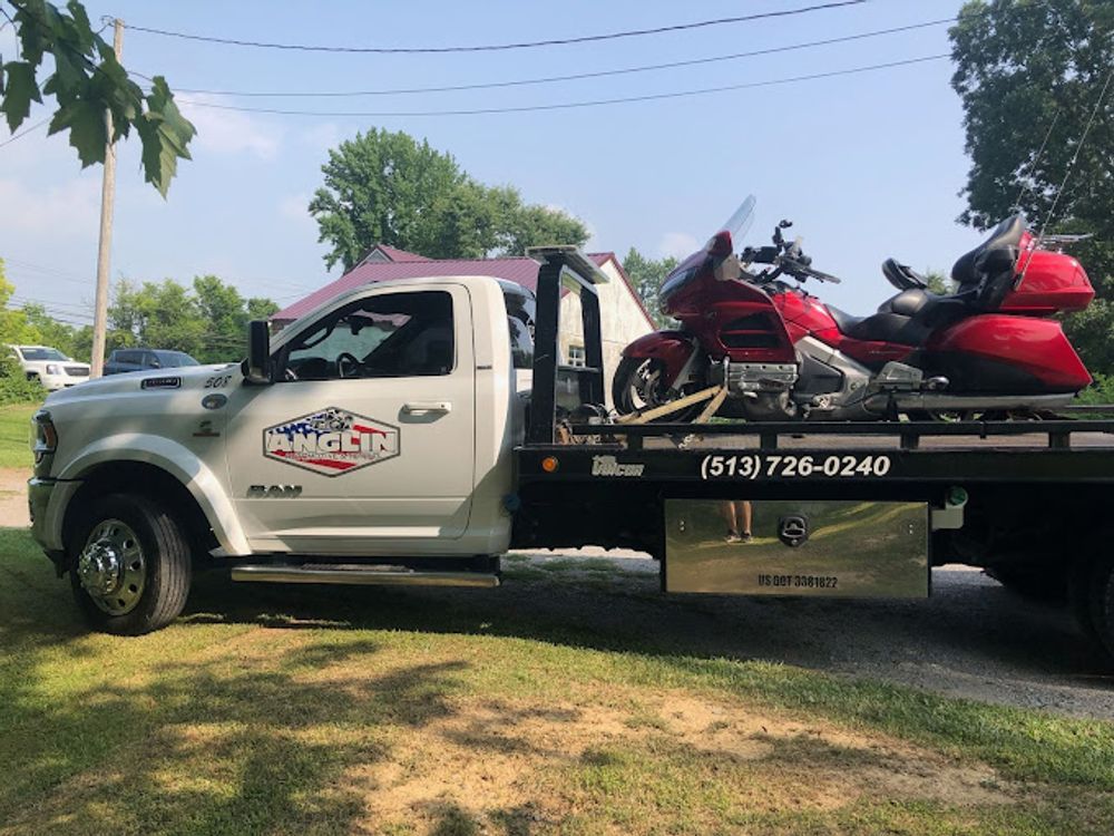 White tow truck with red motorcycle on the flatbed, in a grassy area with trees and a cloudy sky.