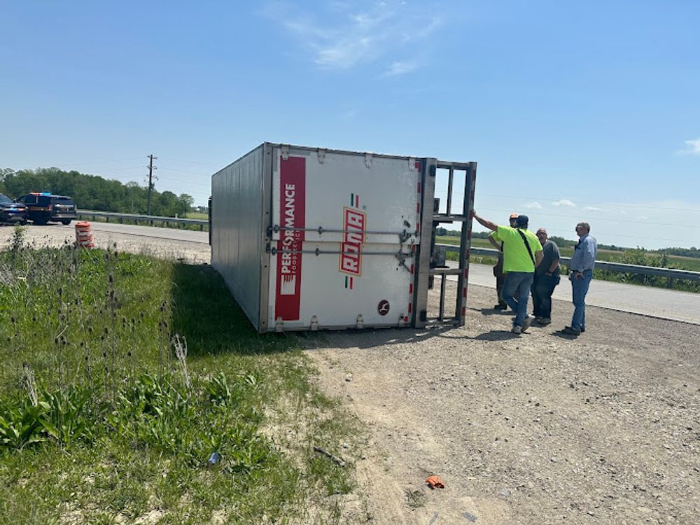 A semi-truck trailer overturned on its side near a road with people assessing the situation.