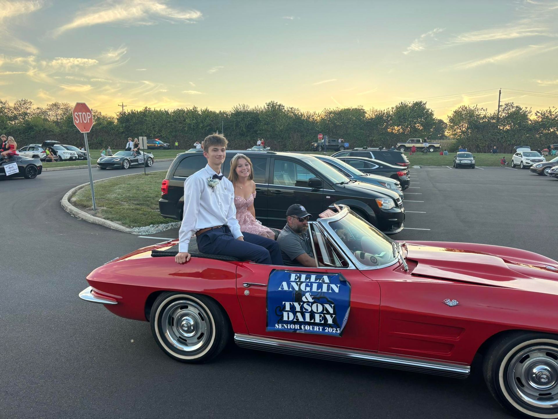 Two teens in formal wear seated on a red classic car in a parking lot. Sign reads: Ally Anselin & Tyson Daly.