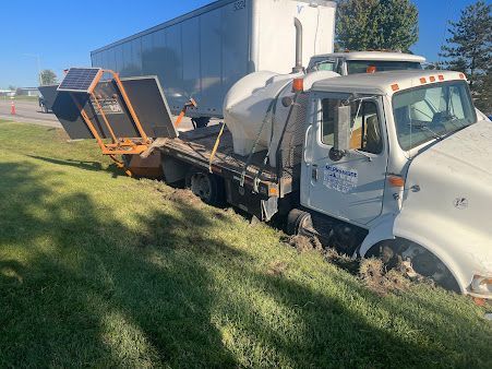 White truck with orange attachments stuck in grass next to a road, trailer in background.