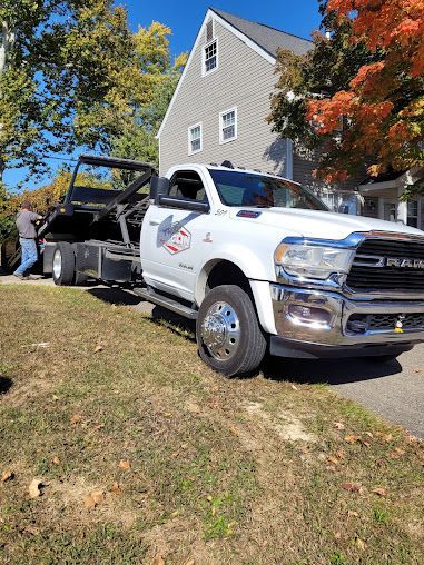 White tow truck with empty bed parked on grass, man standing next to it in front of a house.
