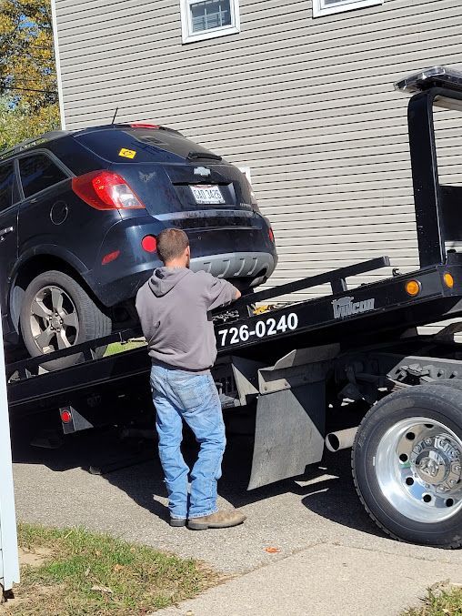 Man loading a dark-colored SUV onto a tow truck. Truck is on a driveway next to a building.