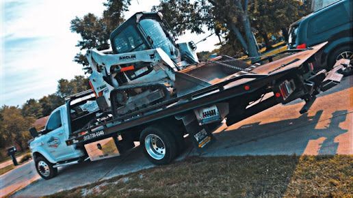 White pickup truck transporting a Bobcat mini-excavator on its flatbed trailer on a residential street.