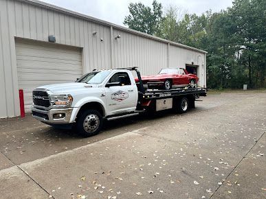 White tow truck with a red car on the flatbed, parked in front of a tan building.