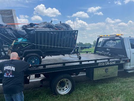 A tow truck carrying a damaged dump truck; a man in a black shirt observes.