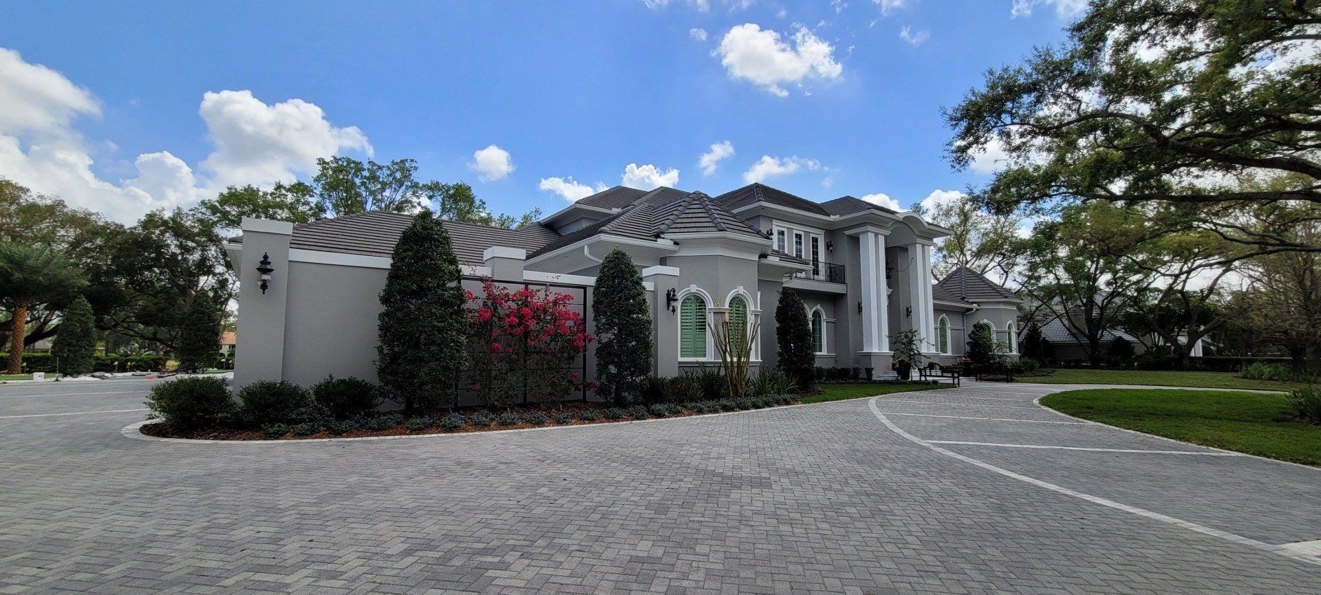 Gray house with a long cobblestone driveway, under a blue sky with clouds.