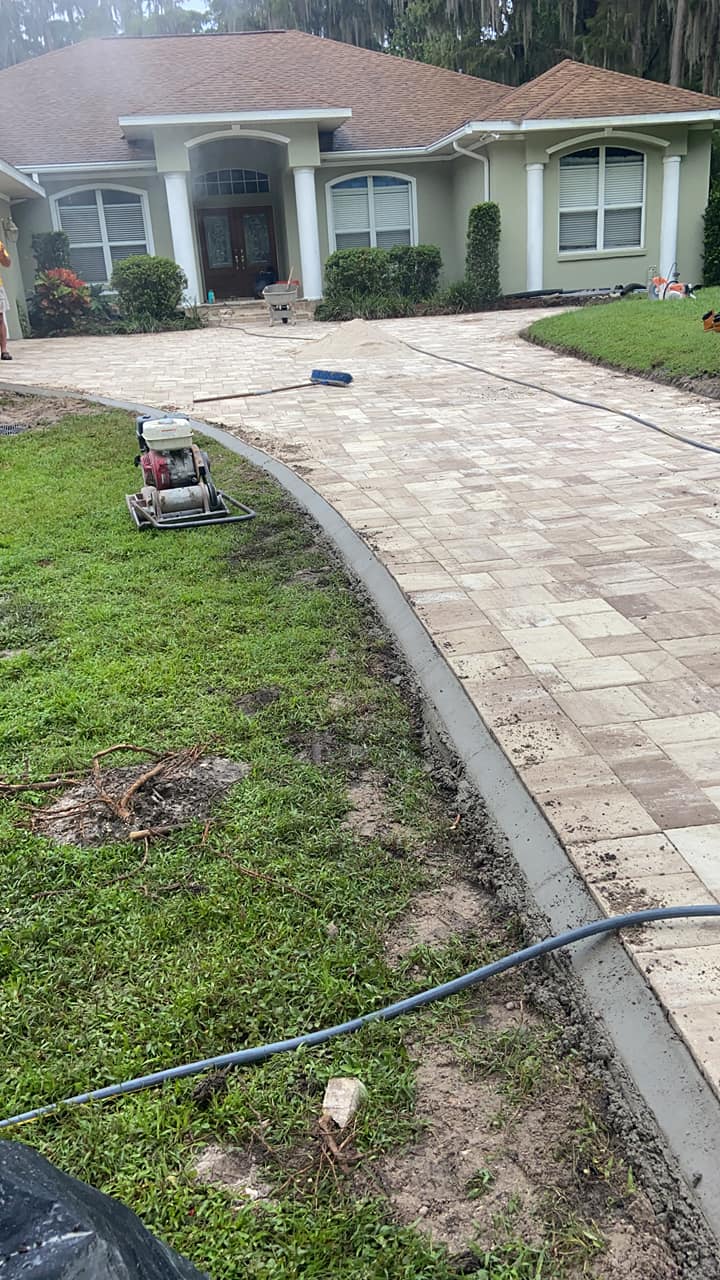 Driveway paving in progress. Beige pavers, concrete edging, house with green siding.