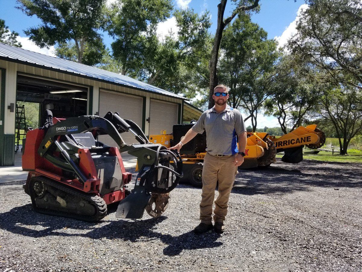Man stands with a stump grinder in a gravel yard. Other equipment and a building are in the background.
