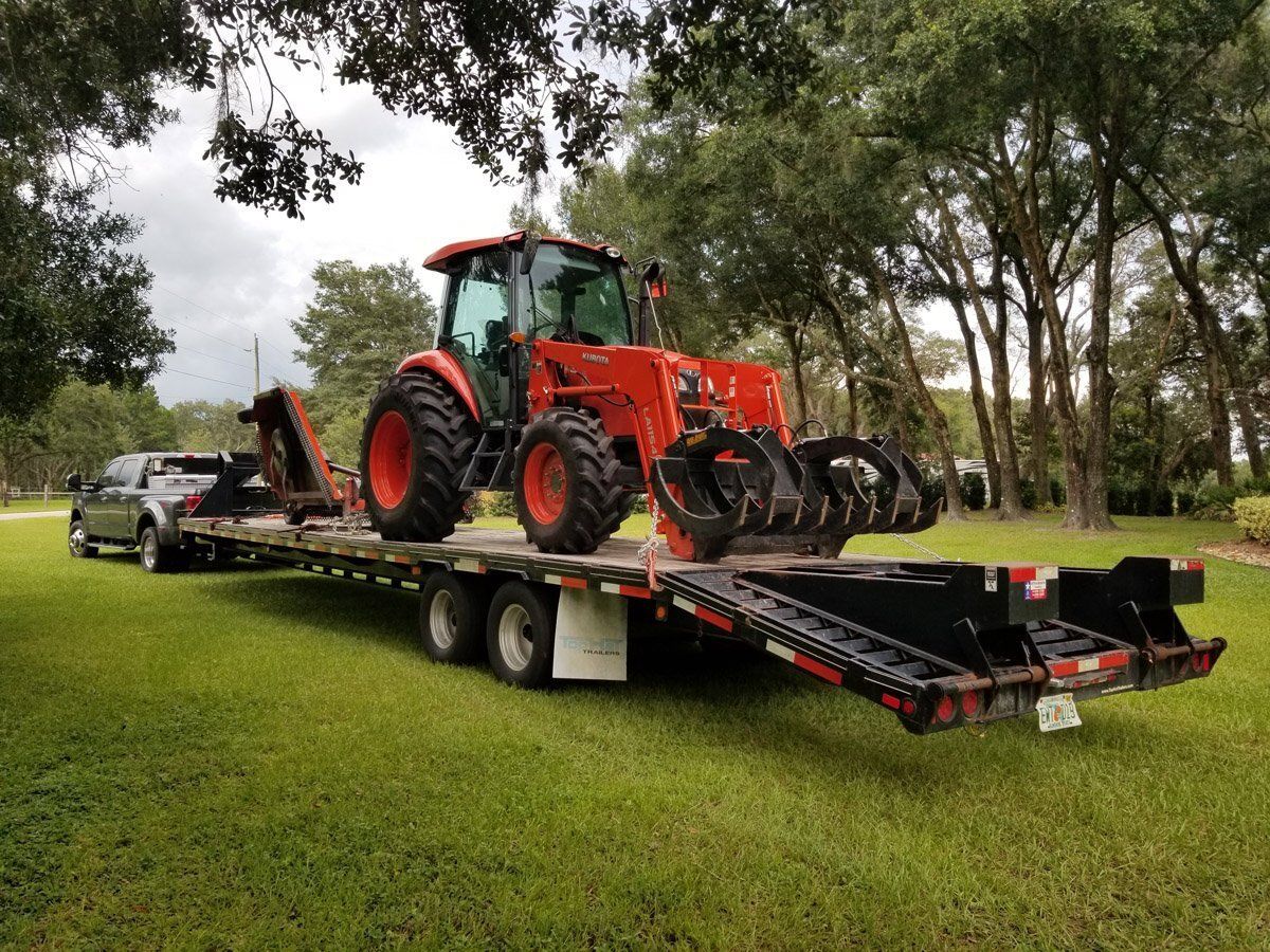 Orange tractor on a trailer, being towed by a silver pickup truck, on green grass.