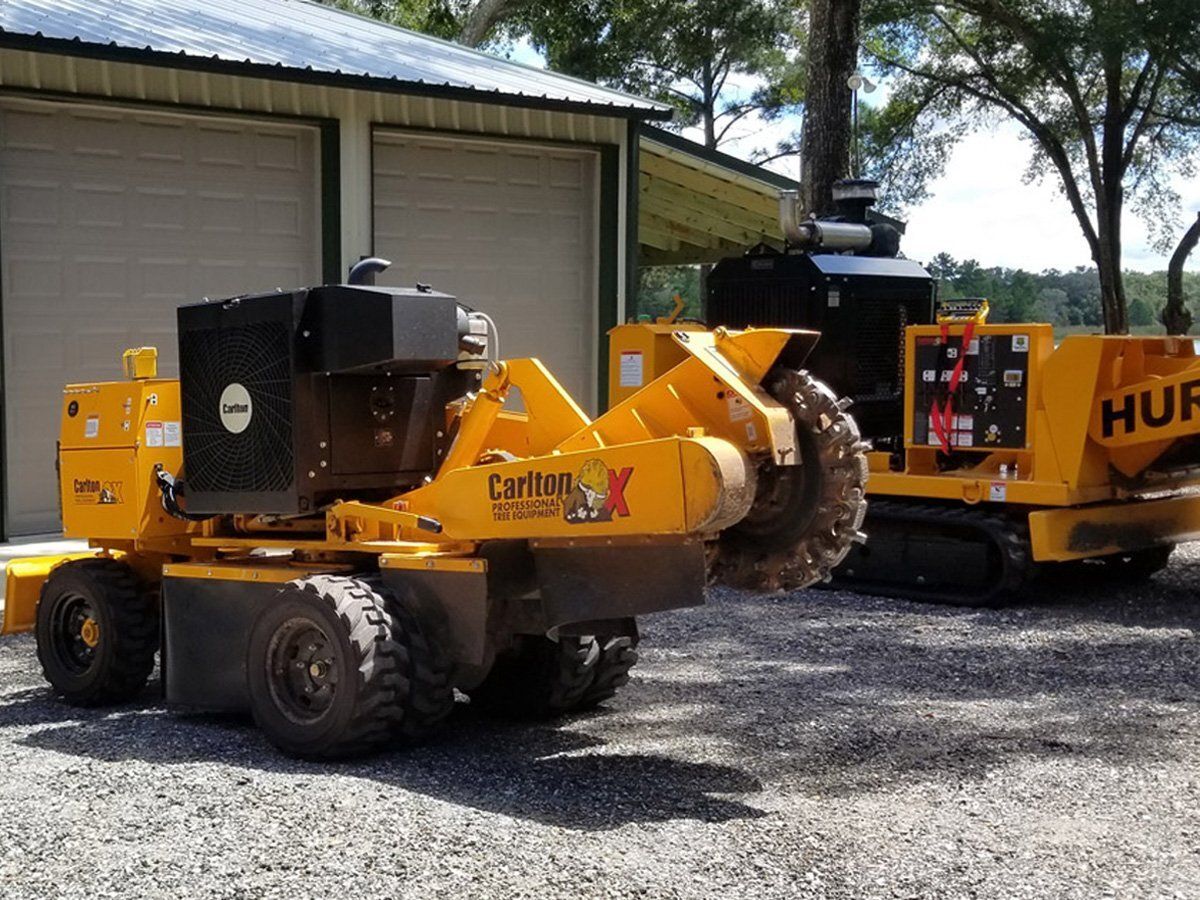 Yellow and black stump grinder on gravel, another in the background near a garage.
