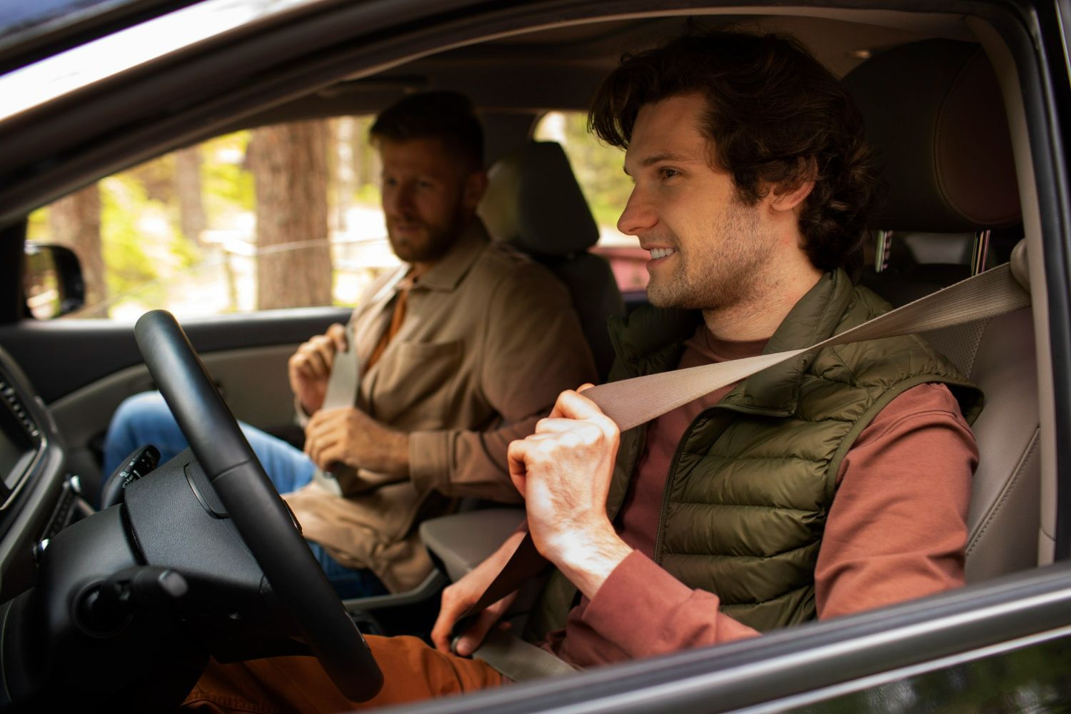 Two men in a car, fastening seatbelts. One is driving, the other in the passenger seat. Forest background.