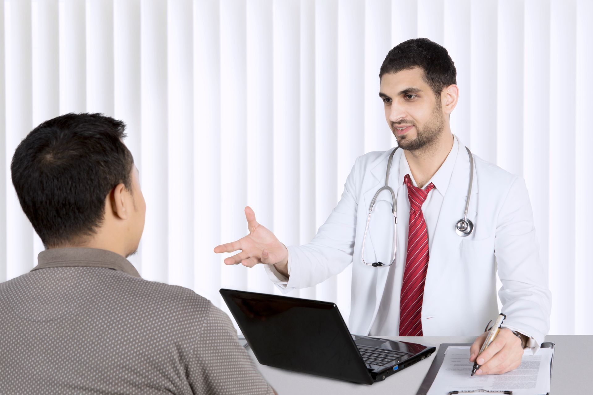 A doctor is talking to a patient while sitting at a desk with a laptop.