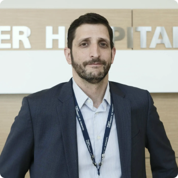 Man in blazer and light blue shirt, standing near a hospital sign. He has a beard and is wearing a lanyard.