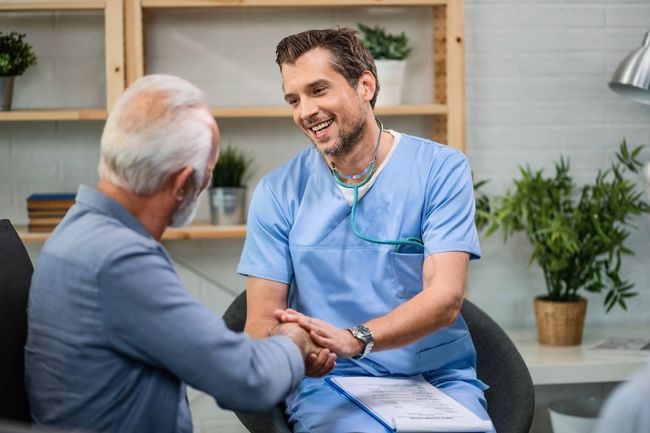 A man in a bathrobe is sitting on a bed talking to a doctor.