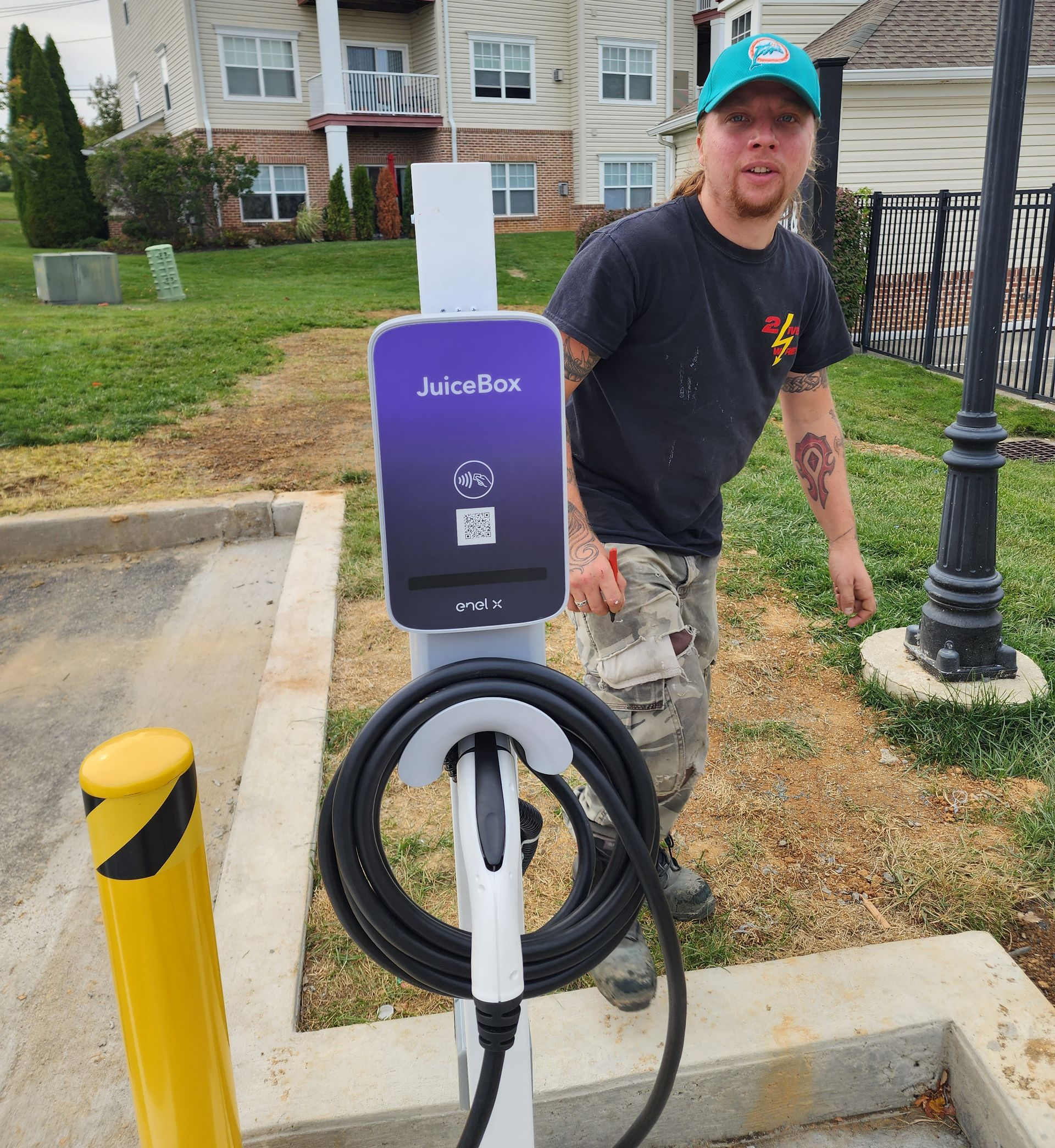 Man standing next to a JuiceBox electric vehicle charger. Outdoor setting with grass and an apartment building.