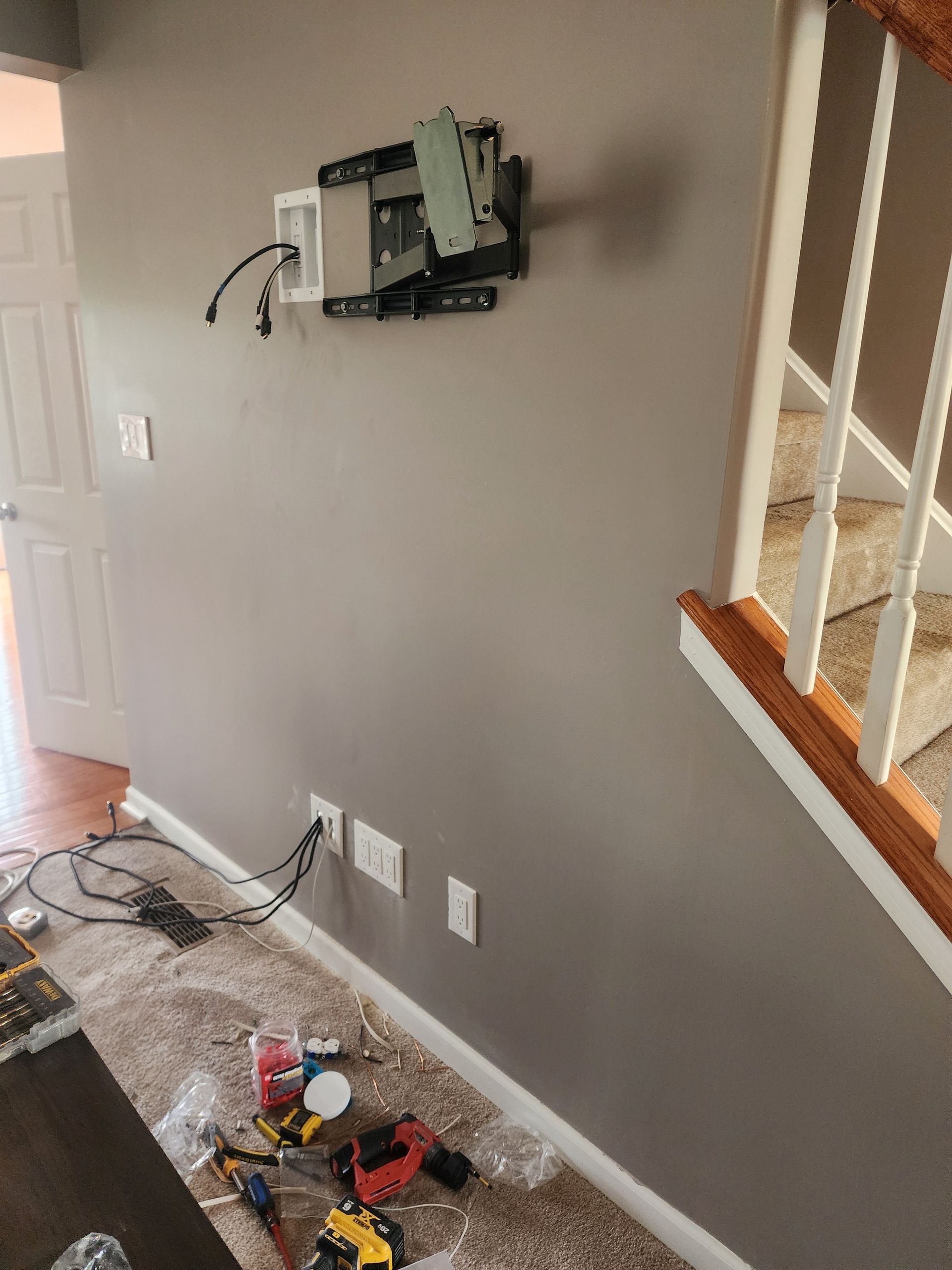 Gray wall with TV mount, wiring, electrical outlets, and tools on the floor. Staircase on right.