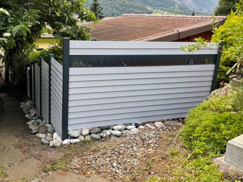 Une clôture blanche est entourée de rochers et d'arbres devant une maison.