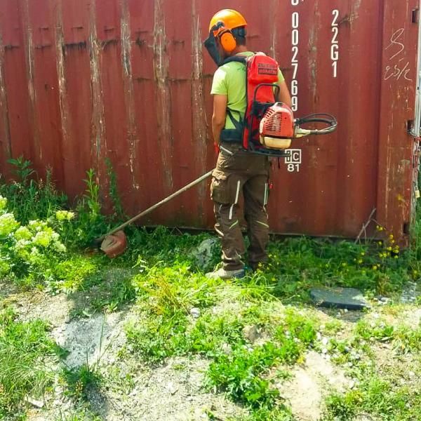Un homme avec un sac à dos coupe l'herbe devant un conteneur d'expédition rouge