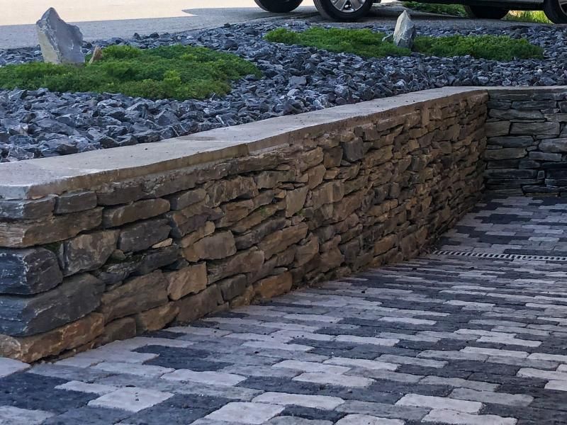 Un mur de pierre entoure une allée en briques avec une voiture garée en arrière-plan.