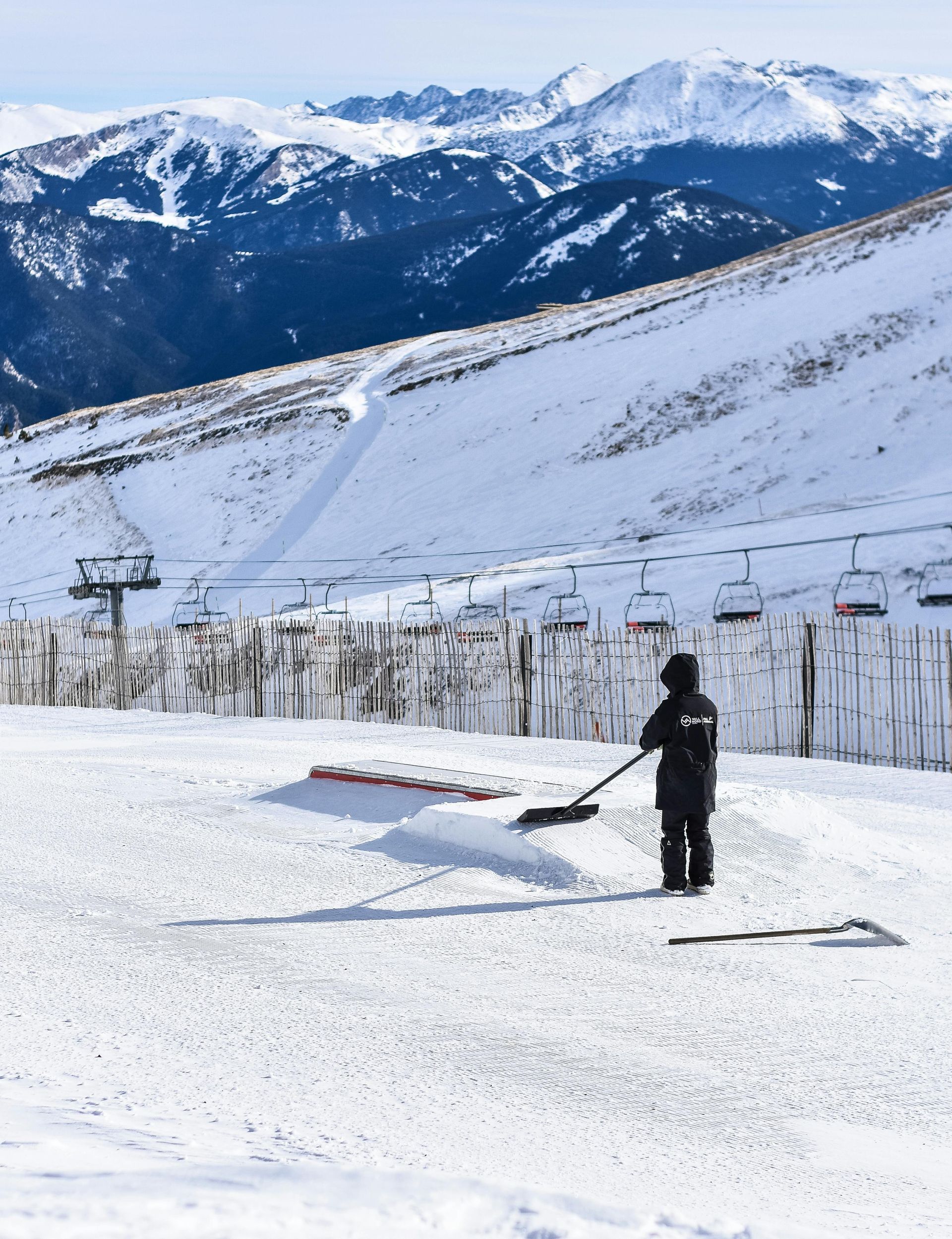 Une personne pellette la neige sur une piste de ski avec des montagnes en arrière-plan.