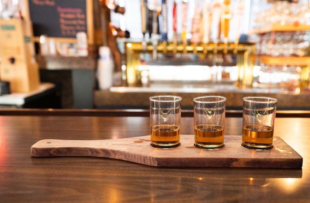 Three shot glasses of whiskey are sitting on a wooden cutting board on a bar.