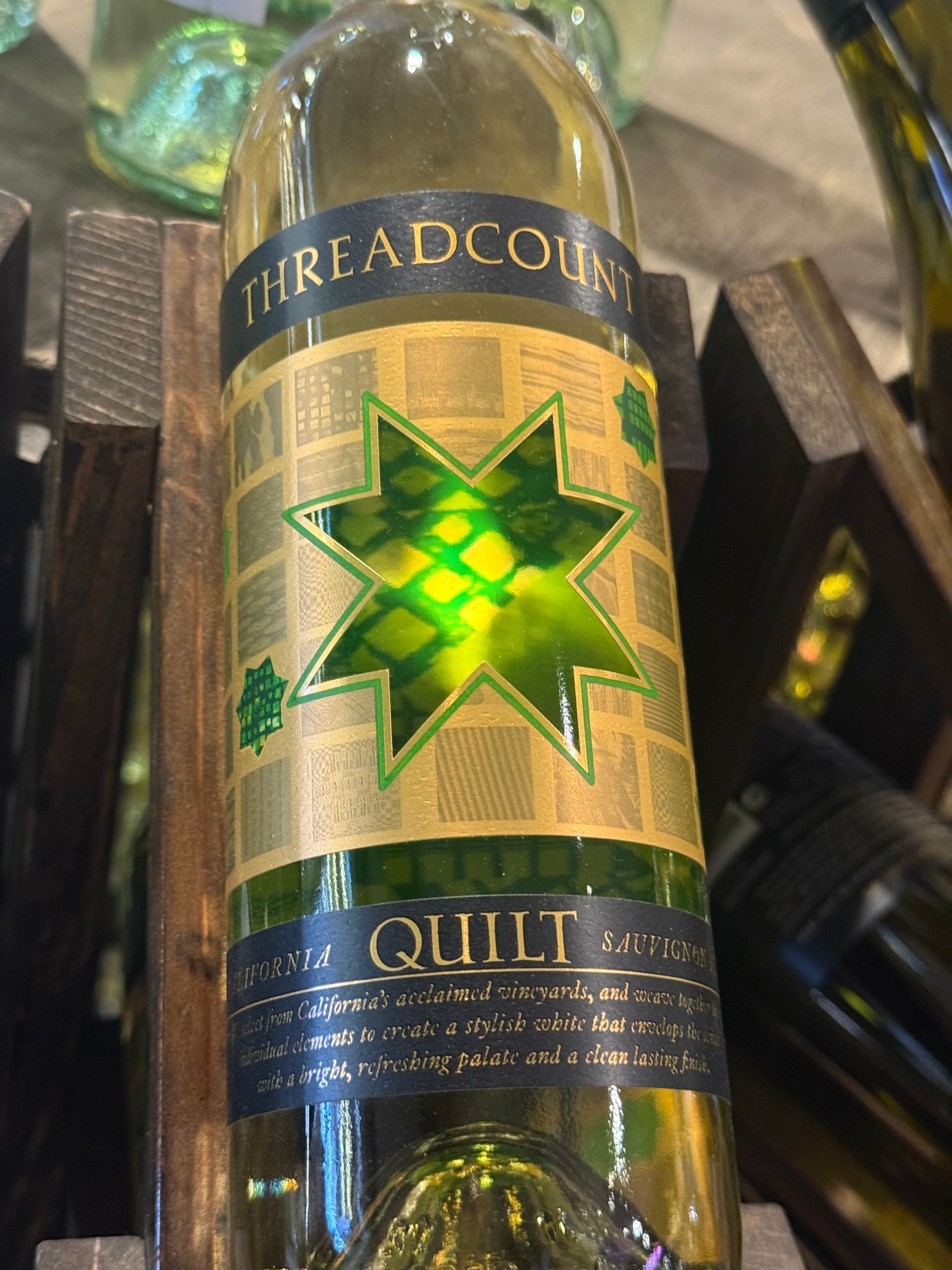 Bottle of Threadcount Quilt wine, gold label with green starburst design, in a wooden wine rack.