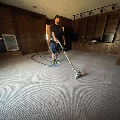 Man vacuuming a carpeted room. The man is using an upright vacuum cleaner with a hose attached, and the room has wood paneling.