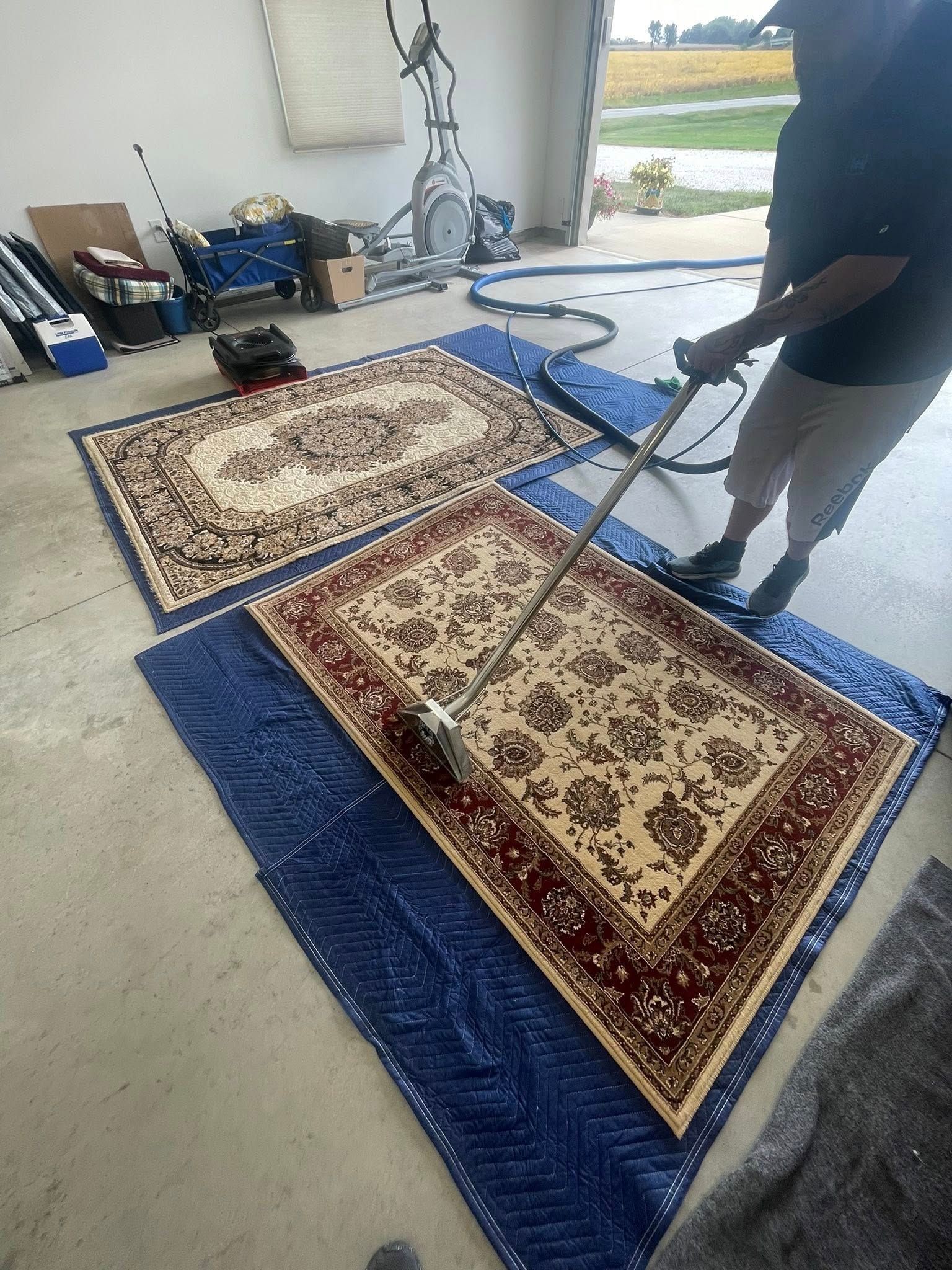 Person cleaning a rug with a machine outdoors on a blue mat; another rug nearby.