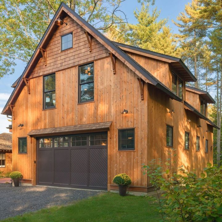 Two-story wooden barn-style building with a gray garage door, surrounded by trees and grass.