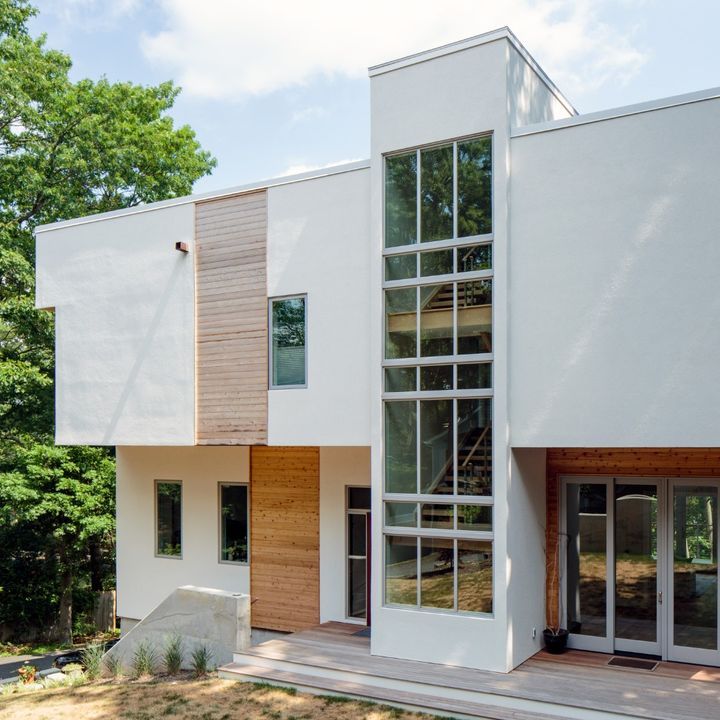 Modern white house with glass windows and wooden accents, set amidst green trees.