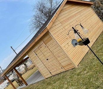 Wooden shed with slanted roof and carport; a bird feeder is in front of it.