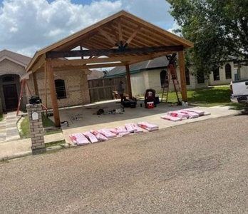Carport construction on a driveway. Beige brick house, wood beams, pink insulation.