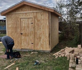 Man building a wooden shed outdoors. Bricks and tools are on the ground nearby.