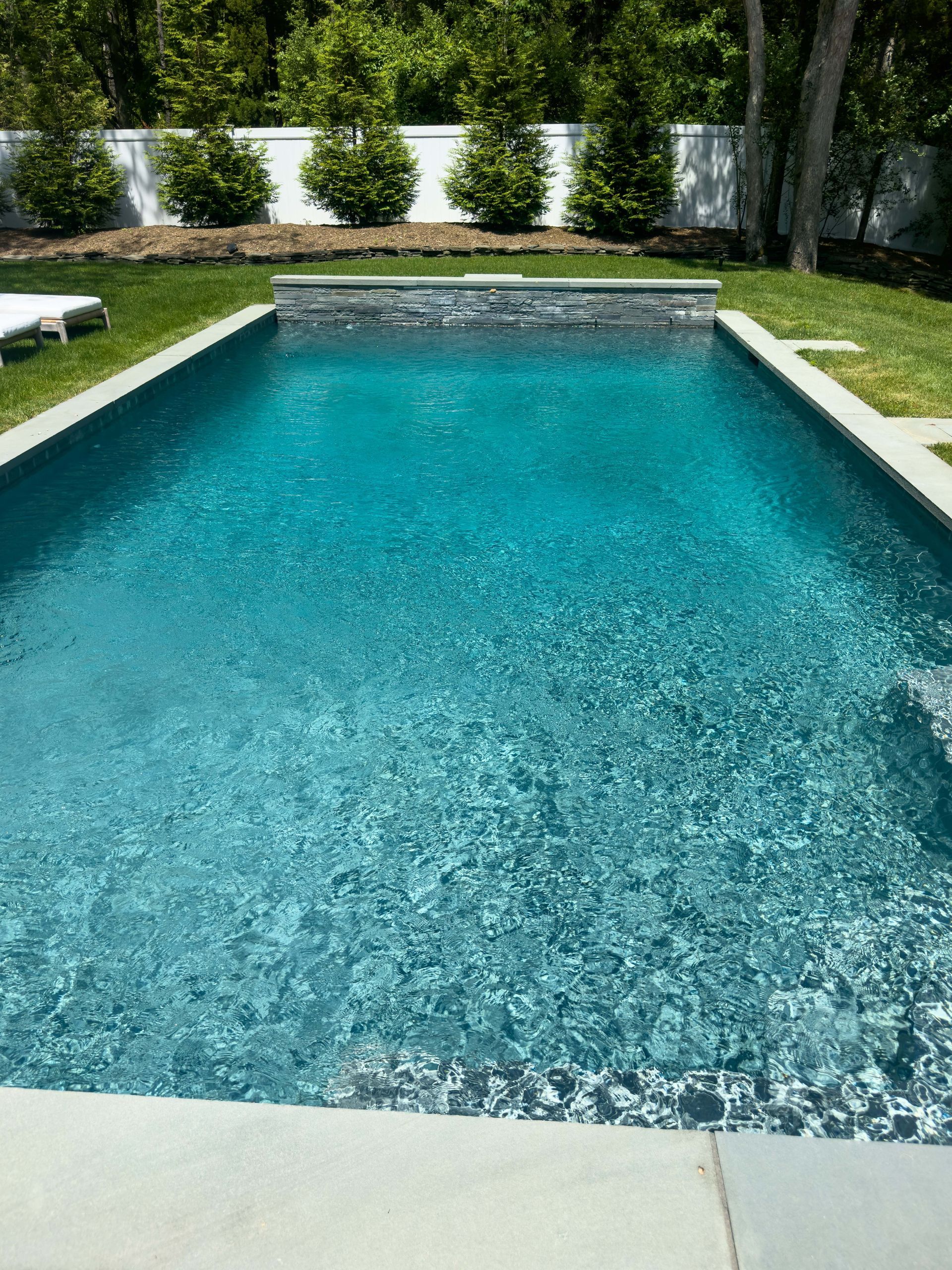 Rectangular swimming pool with teal water surrounded by green grass and a white wall, sunny outdoor setting.