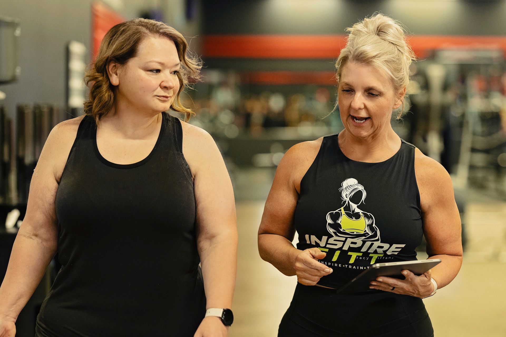 Two women are standing next to each other in a gym looking at a tablet.