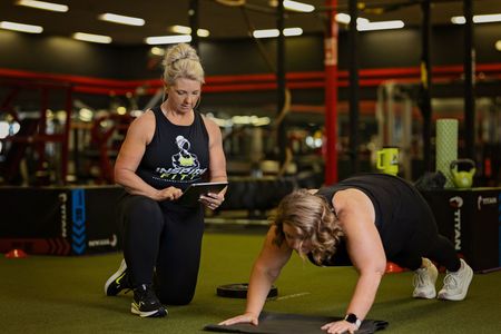 A woman is doing push ups in a gym while another woman looks at a tablet.