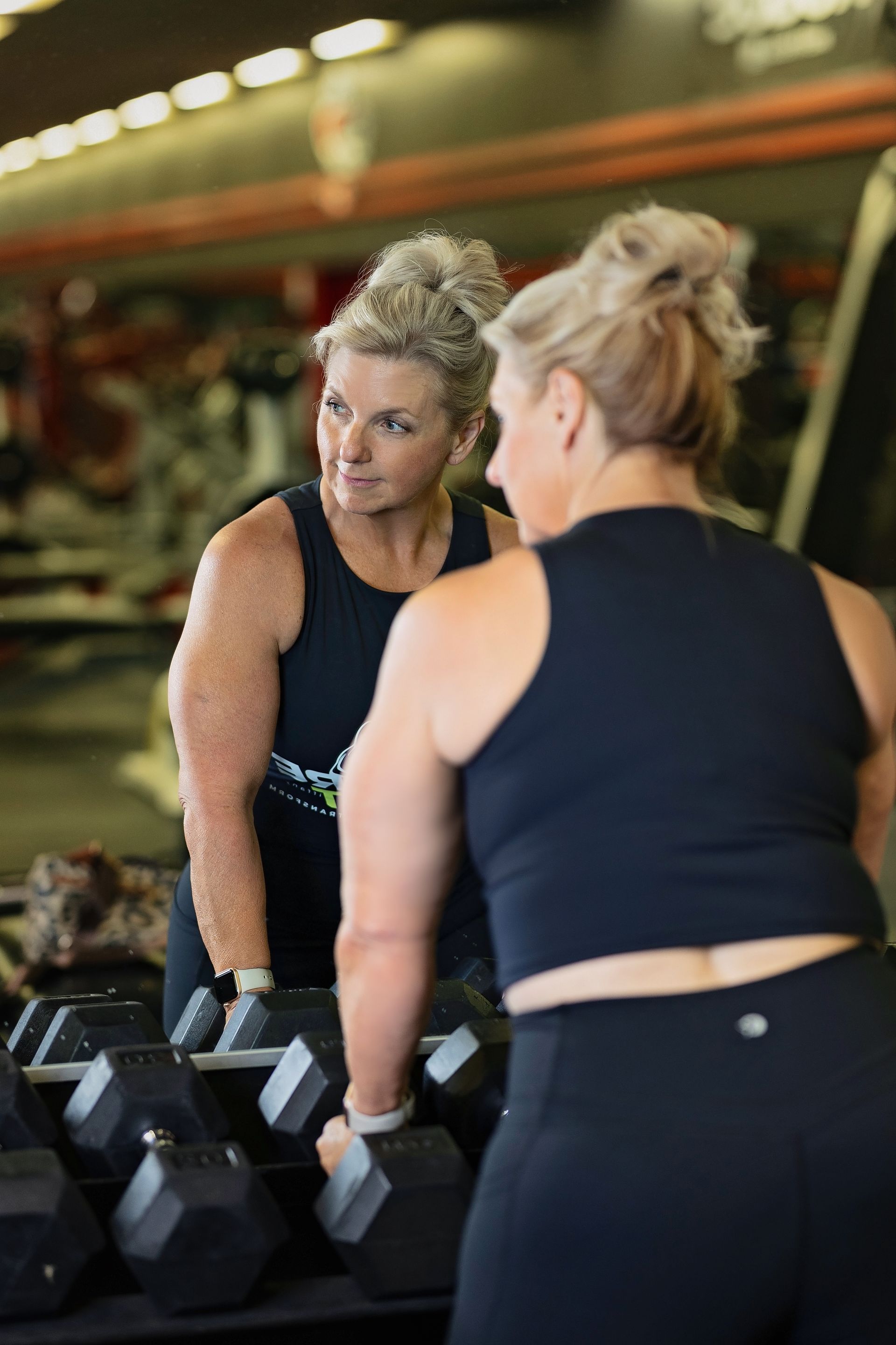 A woman is standing in front of a mirror in a gym holding dumbbells.