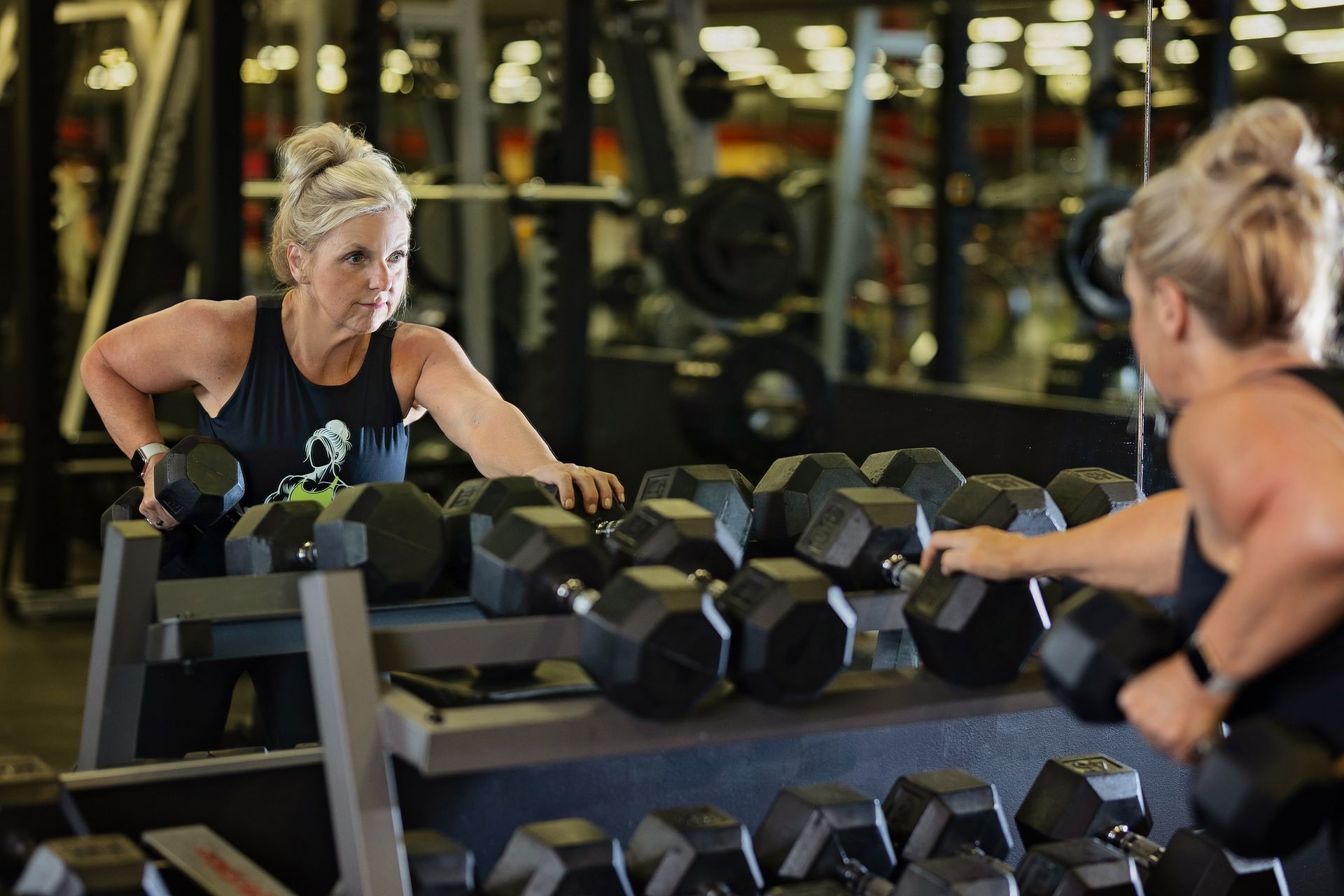 A woman is lifting dumbbells in a gym and looking at herself in the mirror.