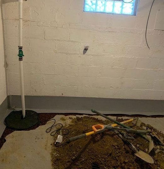 Basement with sump pump, small window, tools in dirt. White walls, grey trim, damp floor.