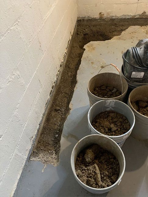 Basement corner with a trench dug along the wall; buckets of dirt sit nearby. Concrete walls and floor.